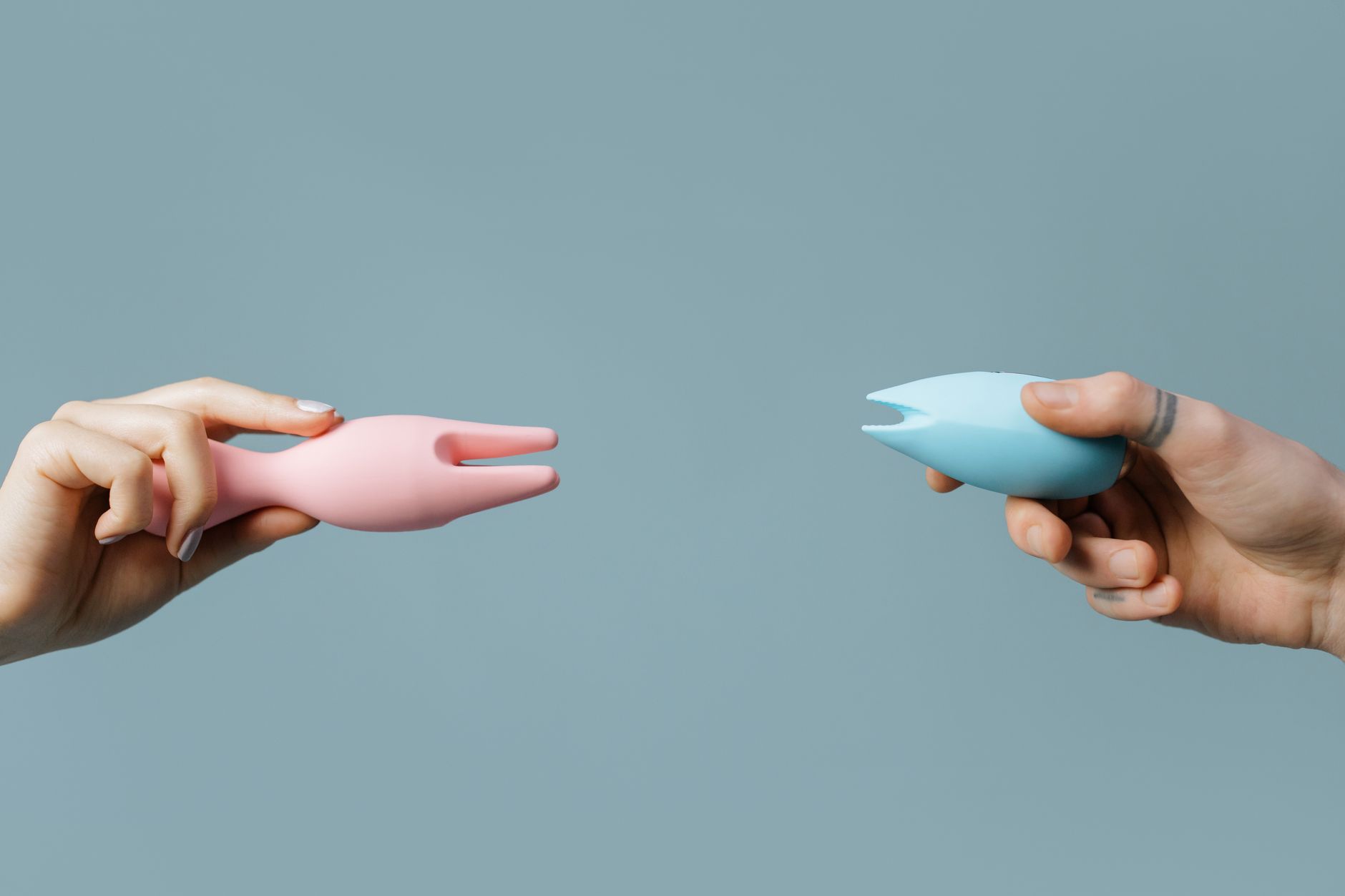 Main tenant un vibromasseur rose et bleu contre un fond pastel minimaliste.