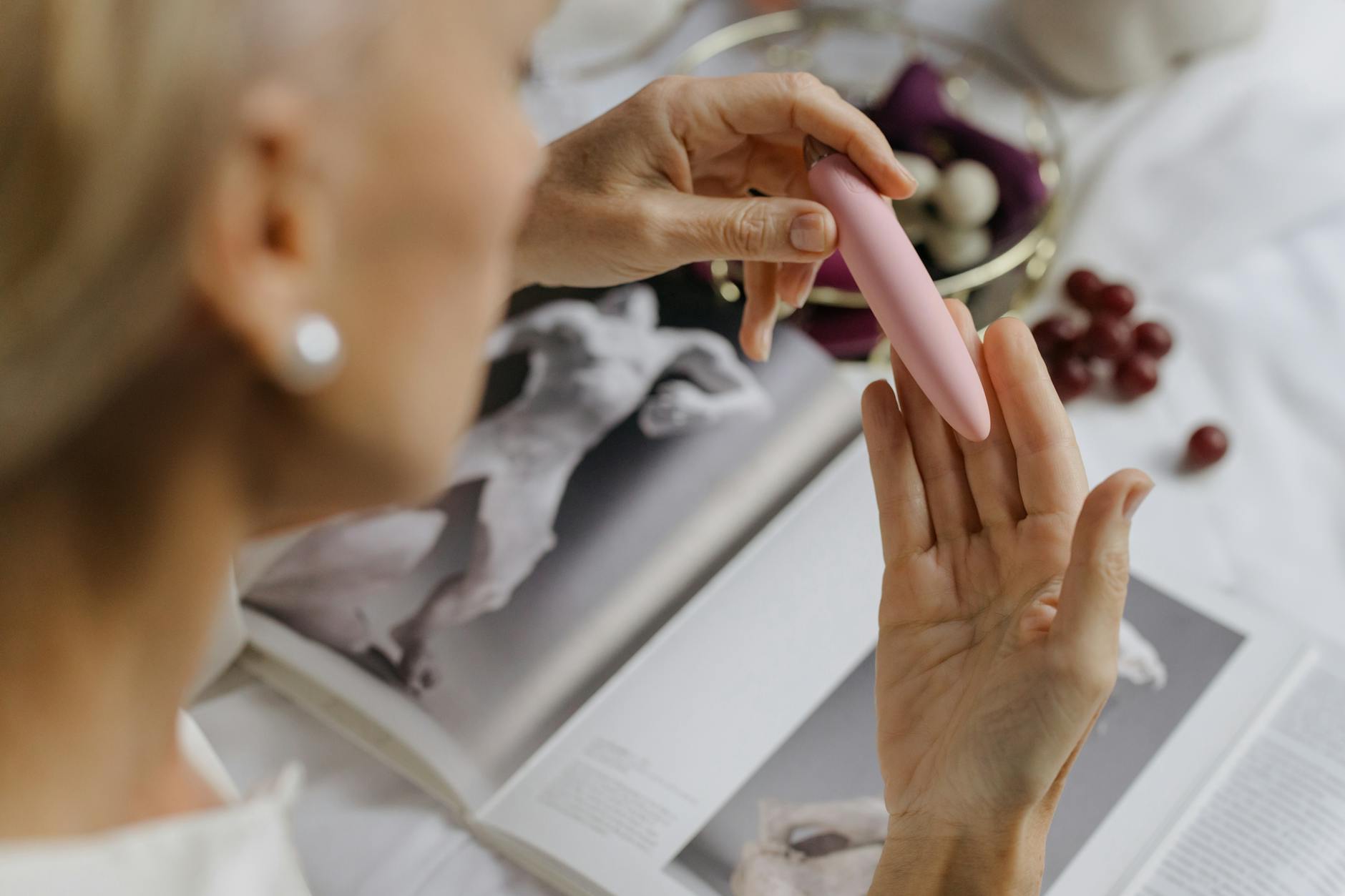 Gros plan d'une femme tenant un vibromasseur rose, avec des livres d'art en arrière-plan, mettant l'accent sur les thèmes intimes et personnels.