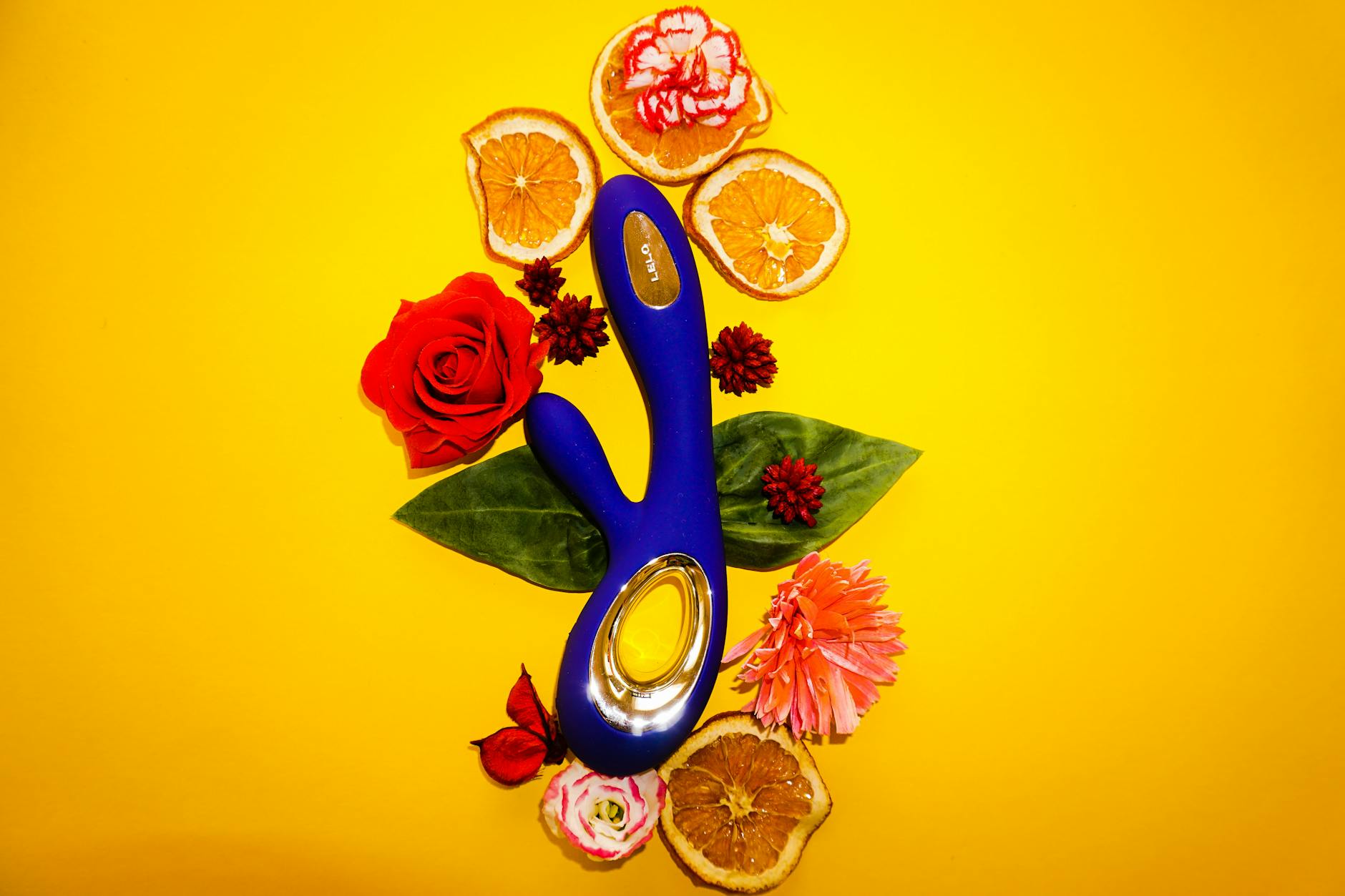 Un vibromasseur bleu entouré de fleurs et de tranches d'orange sur un fond jaune vif